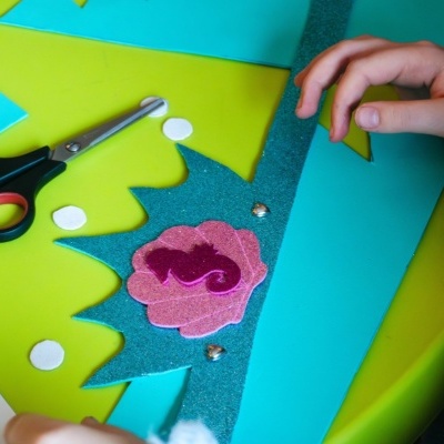 Coroa de EVA azul com decoração de flor rosa e lantejoulas sobre mesa verde com tesoura e pedaços de papel