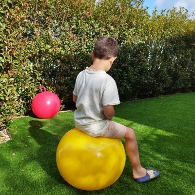 Menino sentado numa bola amarela, bola vermelha ao fundo, relvado artificial com sebe verde