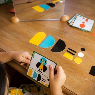 Peças coloridas de cartão a formar um pássaro em tabuleiro de madeira com criança a segurar uma carta.