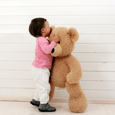 Peluche grande de urso castanho claro ao lado de criança em ambiente interior com parede branca