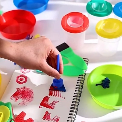 Mão a usar carimbo azul num caderno com recipientes coloridos e tinta em volta