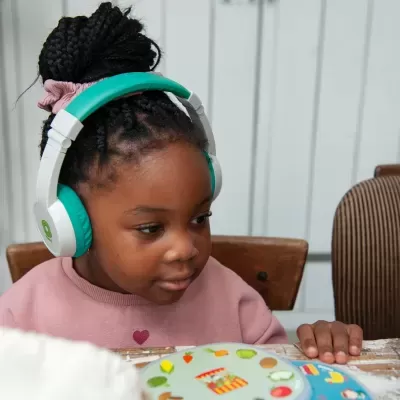 Criança com auscultadores brancos e verdes usando casaco rosa sentada numa mesa com objetos coloridos.