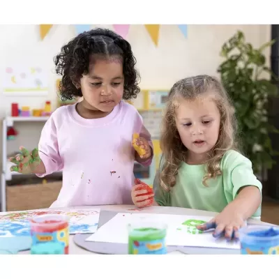 Duas crianças pequenas a pintar com tinta nas mãos em mesa com papéis e tintas.