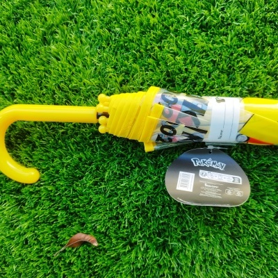 Guarda-chuva amarelo fechado com etiqueta Pokémon sobre relva verde.