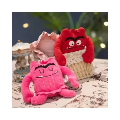Bonecos de pelúcia monstros cor-de-rosa e vermelho sobre mesa com toalha estampada e cesto de vime.