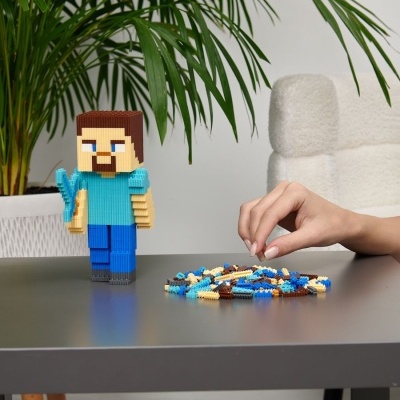 Boneco de bloco de montar como figura quadrada com picareta azul e peças soltas numa mesa cinzenta, com planta verde e cadeira branca ao fundo