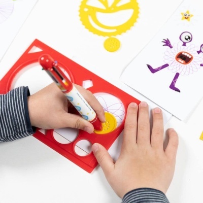 Mãos de criança a desenhar com caneta num stencil vermelho, próximo de um desenho de monstro roxo e amarelo