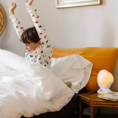 Criança a espreguiçar-se na cama branca com almofada amarela e luz de mesa redonda