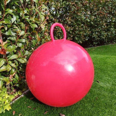 Bola de exercício infantil vermelha com pega sobre relva artificial e arbusto verde