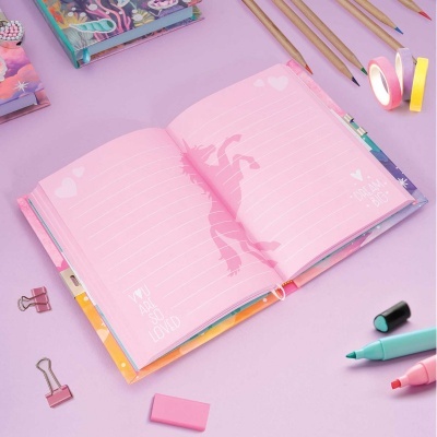 Caderno rosa aberto com unicórnio e texto, lápis e objetos de papelaria sobre fundo rosa