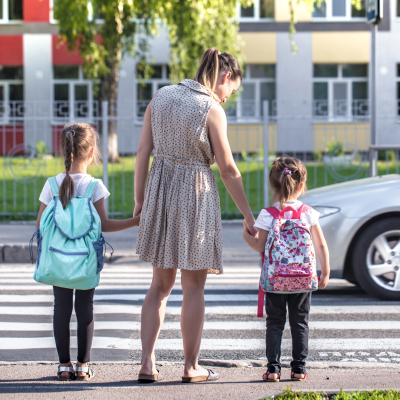 Dois crianças com mochilas e mulher a atravessarem passadeira na cidade com carro e edifícios coloridos ao fundo