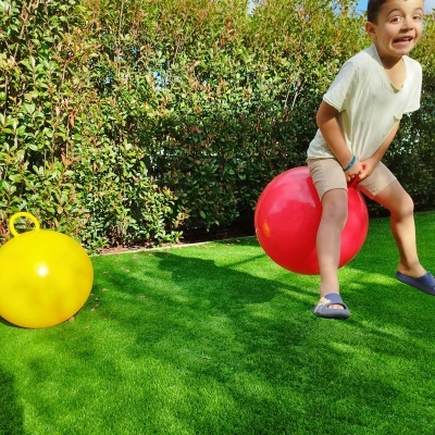 Criança a saltar numa bola insuflável vermelha num relvado artificial com bola amarela ao lado e vegetação atrás
