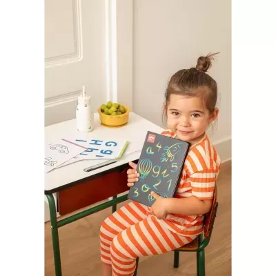 Criança com pijama laranja às riscas segura quadro com desenhos coloridos sentada à mesa branca com papel e fruta.