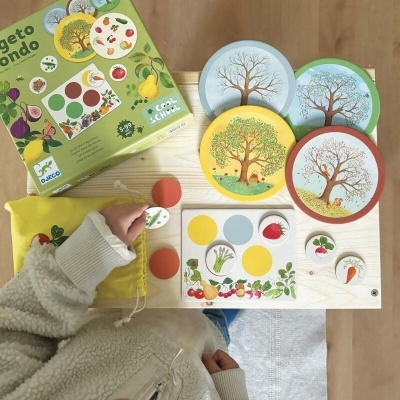Jogo educativo com peças redondas de madeira, ilustrações de árvores e frutos, saco amarelo e caixa verde