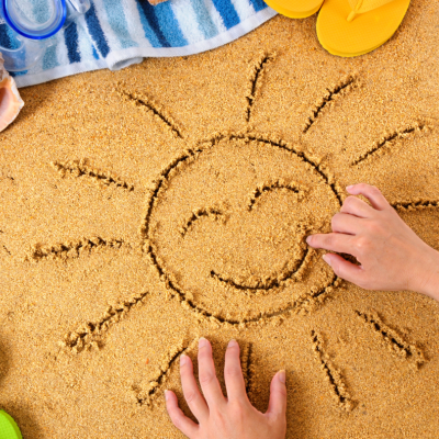 Mãos desenhando sol sorridente na areia com acessórios de praia ao fundo