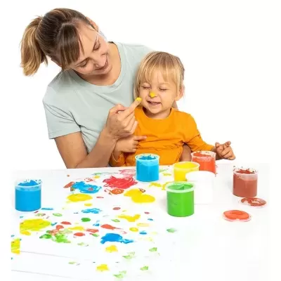 Mulher e criança a pintar com tinta colorida numa mesa branca com frascos de tinta