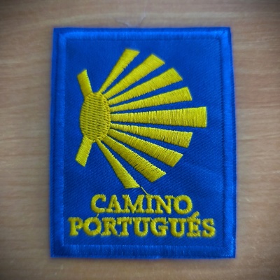 Emblema bordado azul e amarelo com texto CAMINO PORTUGUÊS sobre superfície de madeira