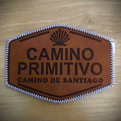 Etiqueta de couro castanho com texto CAMINO PRIMITIVO e CAMINO DE SANTIAGO e desenho de concha