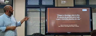 Homem aponta para ecrã digital com texto em português numa sala com janela e persianas.