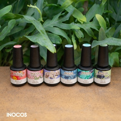 Seis frascos de verniz gel INOCOS com rótulos coloridos e tampas pretas, sobre superfície castanha e folhas verdes ao fundo.