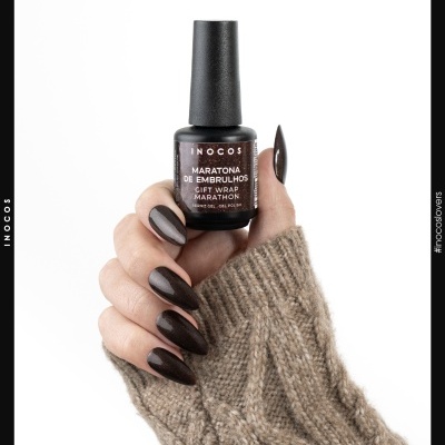 Frasco de verniz gel castanho segurado por mão com unhas pintadas na mesma cor e manga de camisola bege