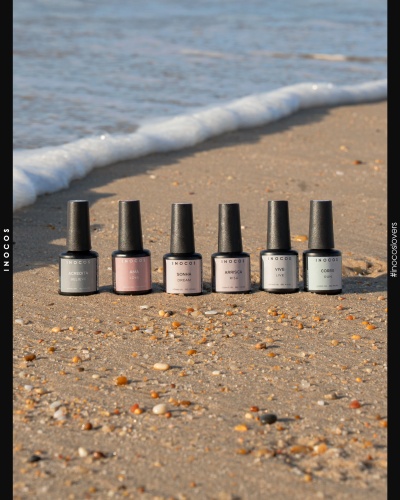 Seis frascos de verniz gel INOCOS em diferentes tons pasteis na areia da praia perto do mar.
