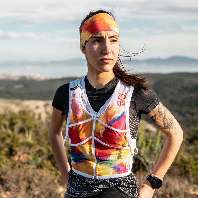 TRAIL RUNNING BACKPACK COLORS Mulher a usar faixa de cabeça e colete coloridos com padrão abstrato num cenário natural