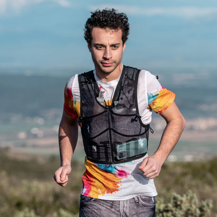 TRAIL RUNNING BACKPACK BLACK JEANS Homem a correr com mochila de hidratação preta e camiseta colorida ao ar livre
