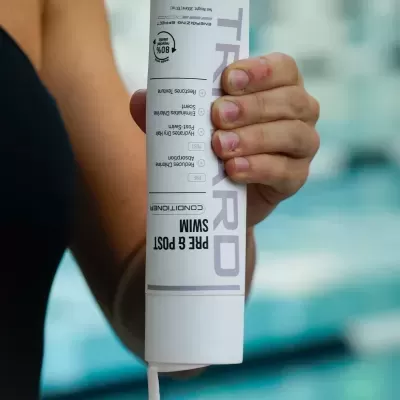 Frasco branco de TRI PRO conditioner PRE E POST SWIM sendo espremer por uma pessoa junto a uma piscina.