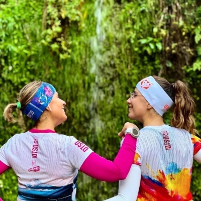 Duas mulheres de costas com roupas desportivas coloridas e faixas na cabeça, em ambiente natural com cascata