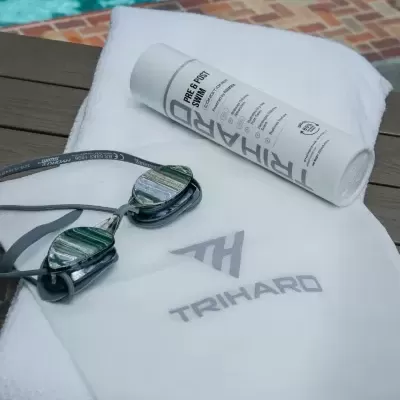 Toalha branca com logótipo TRI HARD, óculos de natação espelhados e frasco branco de condicionador TRI HARD junto a piscina