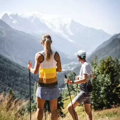 Duas pessoas a caminhar com bastões em trilho de montanha, vista de costas, montanhas nevadas ao fundo