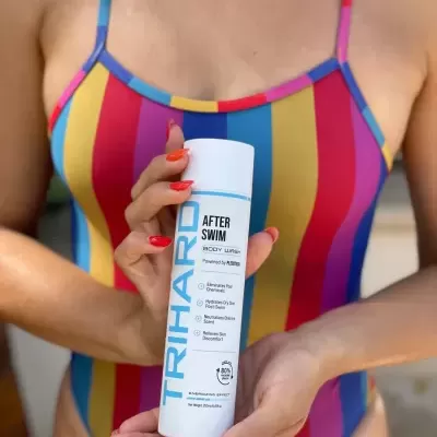 Frasco de loção corporal AFTER SWIM da TRIHAIR com texto azul e preto, segurado por pessoa com fato de banho colorido às riscas.