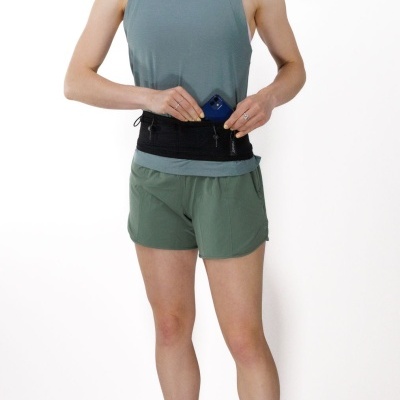 Pessoa com camisola azul esverdeada, calções verdes, meias pretas e tênis brancos, com cinta preta à cintura onde insere um telemóvel azul