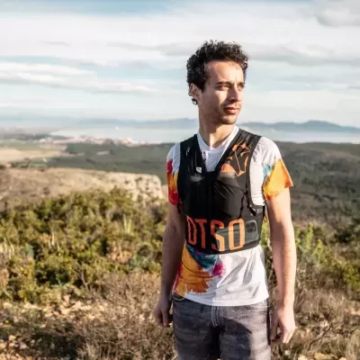 Homem a usar t-shirt colorida e colete preto com letras laranjas ao ar livre com paisagem natural