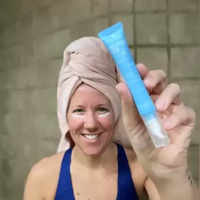 Mulher a segurar tubo azul de produto cosmético com toalha na cabeça