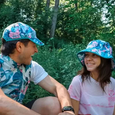 Homem e mulher com bonés e roupa desportiva florais, num cenário natural com muitas plantas verdes.