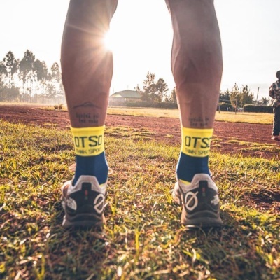 Pernas masculinas com meias azuis e amarelas 'OTSO' e sapatilhas de corrida cinzentas em chão de terra e relva