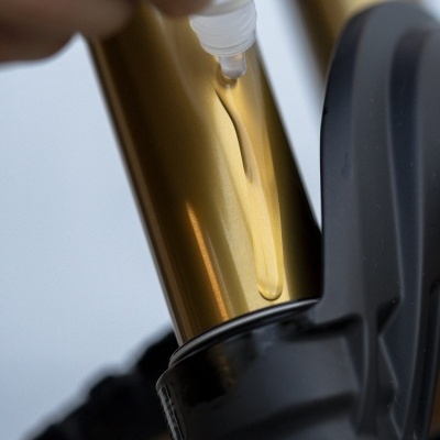 Mão aplicando líquido numa suspensão dourada de bicicleta com inscrição '34 29'