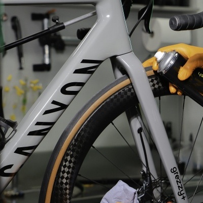 Bicicleta de estrada cinzenta com detalhe da roda e pessoa a aplicar spray com luvas amarelas.