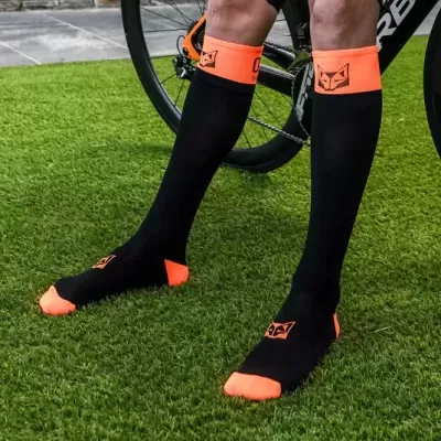 Meias desportivas pretas com detalhes em laranja e logótipo de raposa, usadas junto a bicicleta sobre relva artificial