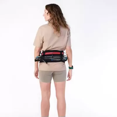 Mulher de costas com t-shirt bege e calções cinza claro, usando cinto com bastões de caminhada.