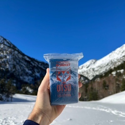 Caixa plástica de manta de emergência OTSO com fundo de montanhas e neve.