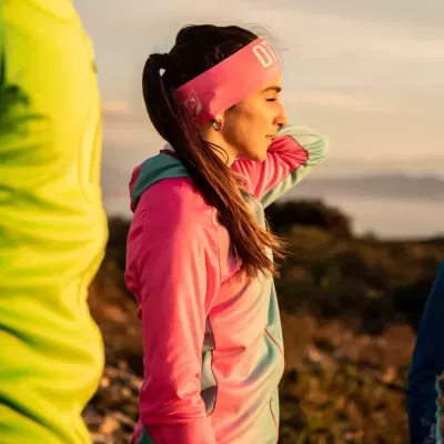 Mulher vestindo roupa desportiva rosa e azul claro, com banda na cabeça rosa e paisagem natural ao fundo