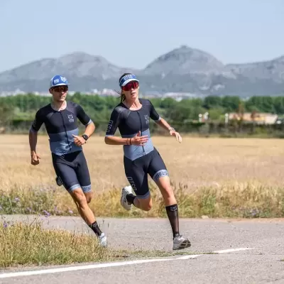 Dois atletas a correr numa estrada em paisagem rural, ambos com equipamento desportivo preto e azul.