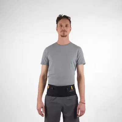 Homem com t-shirt cinza e calças cinza escuro com cinto preto e amarelo em fundo branco.