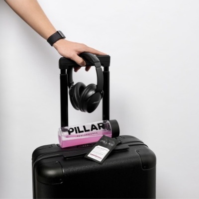 Mala preta com auscultadores, garrafa rosa PILLAR e tubo preto em fundo branco