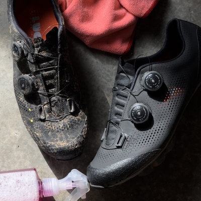 Dois sapatos de ciclismo pretos, um sujo e outro limpo, pano rosa e frasco com spray em chão cinzento