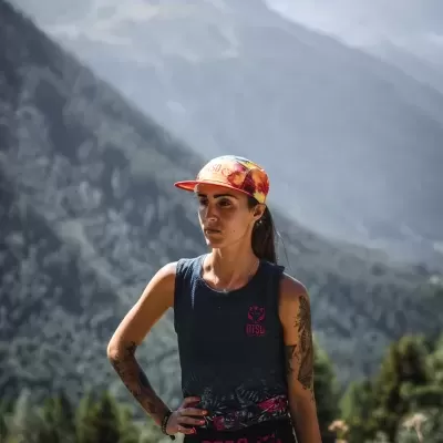 Mulher com camisola preta sem mangas e boné laranja num cenário de montanhas e árvores