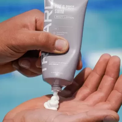 Tubo cinzento claro de creme corporal a ser aplicado na mão com texto branco e fundo azul desfocado